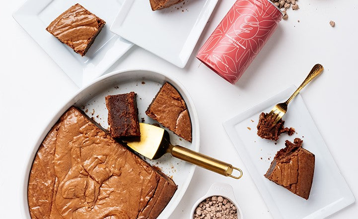 Oval baker with brownies and cut pieces on white rectangle plates and tin of cocoa mix