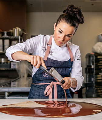 Gnger Elizabeth working with melted chocolate on mable ountertop and commercial kitchen in background.