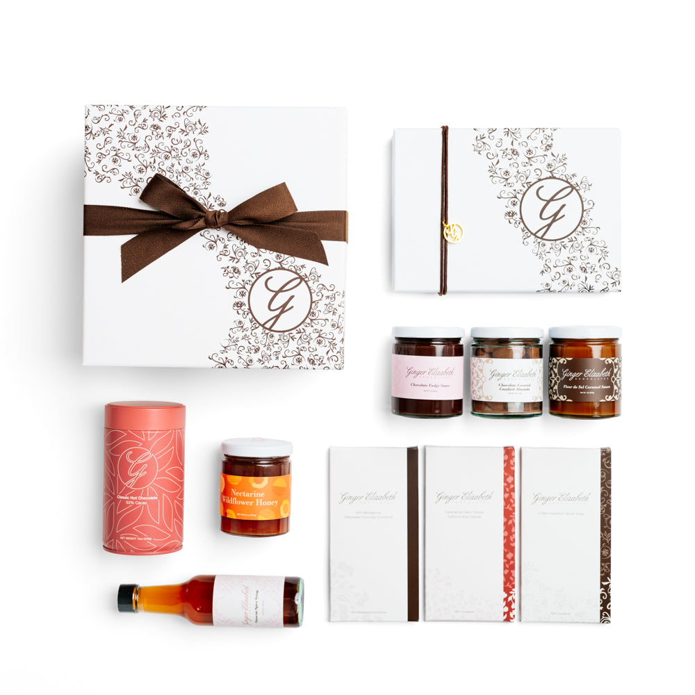 Gift Box shown with 24 piece assorted chocolate bonbon box, 3 chocolate dessert bars, classic hot cocoa tin, chocolate covered almonds, fudge sauce, fleur de sel caramel sauce, artisanal jam and oaxacan spice syrup on white background.