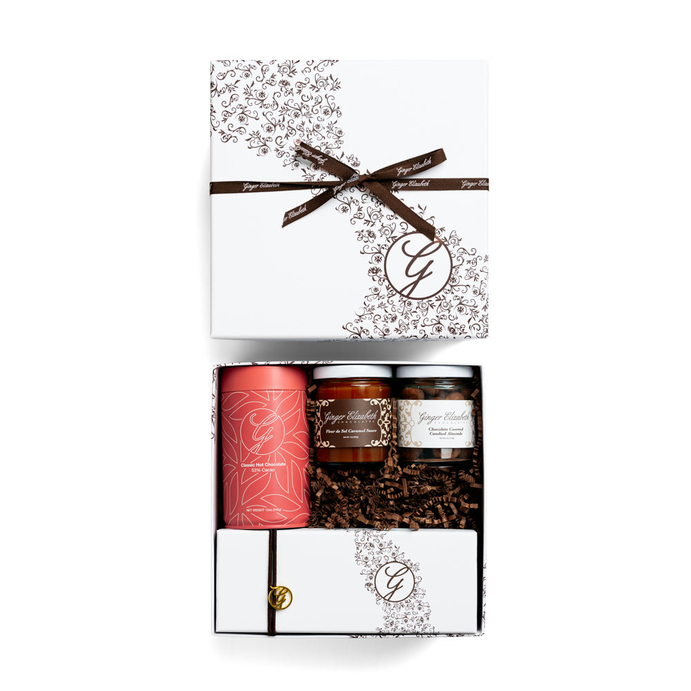 Gift box packaged with 12-piece chocolate box, classic hot chocolate tin and jars of caramel sauce and candied almonds with lid above on white background.