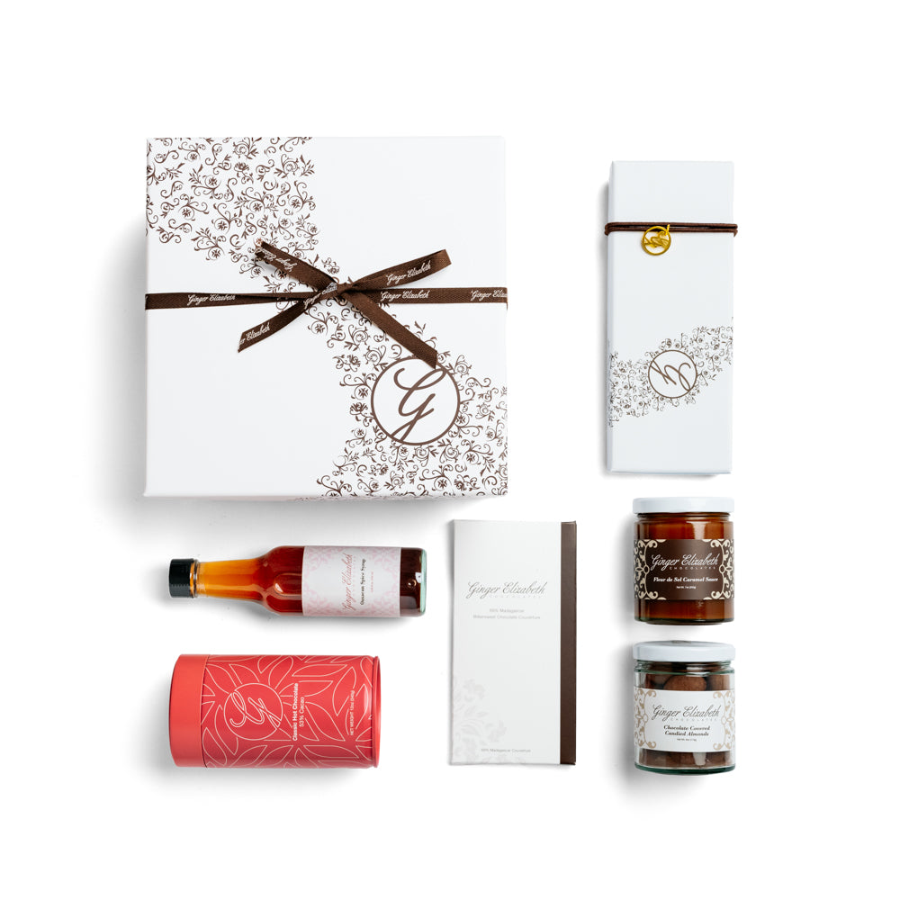 Gift Box surrounded by  12 piece assorted chocolate box, chocolate bar, chocolate covered almonds, classic hot chocolate tin, oaxacan spice syrup and fleur de sel caramel sauce on white background.