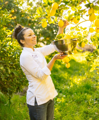 woman in chef's coat and jeans holding a stainless colander and picking lemons