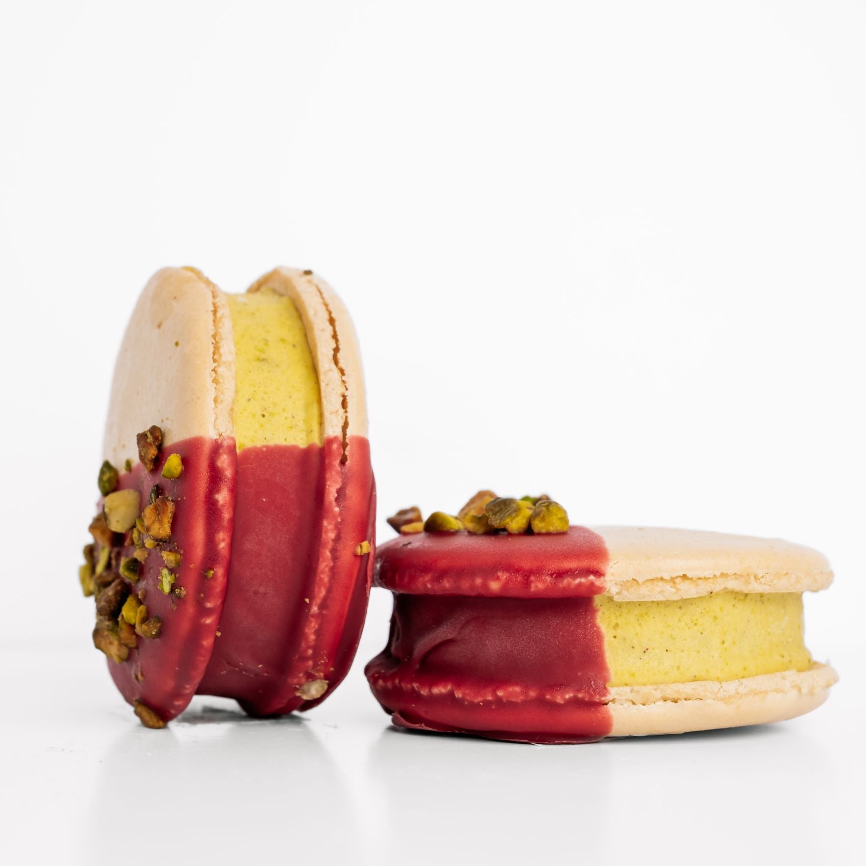 Two macaron ice cream sandwiches dipped in raspberry chocolate with pistachio toppings on a white background