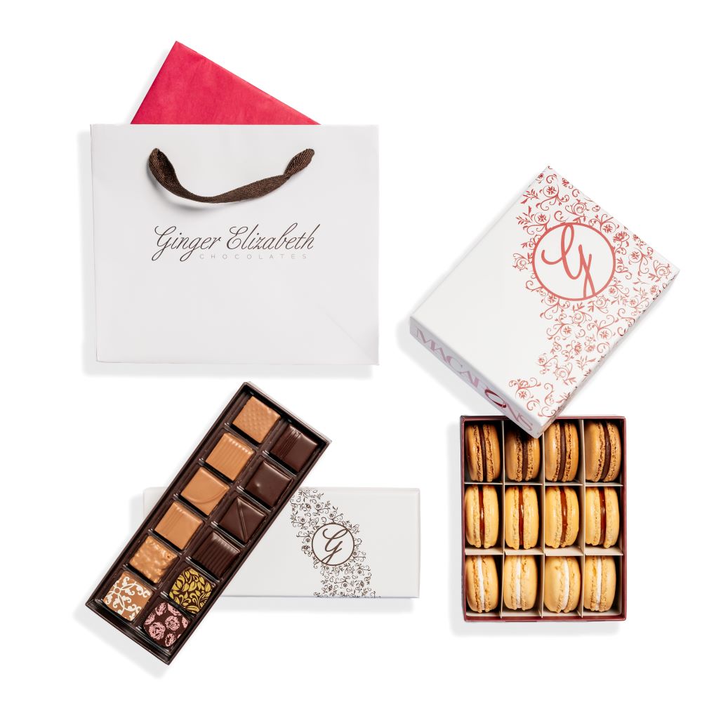 Gift bag with pink tissue, open 12 piece  box of chocolates  and 12 piece macaron box  with lids arranged on white background