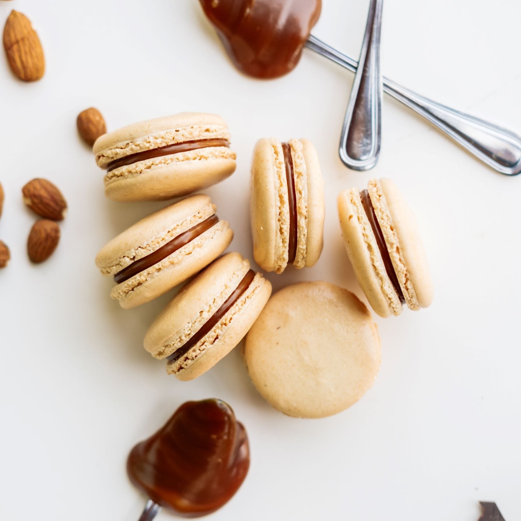 Six Salty Caramel Macarons shown with Caramel filling on spoons  and scattering of fresh roasted almonds on white background