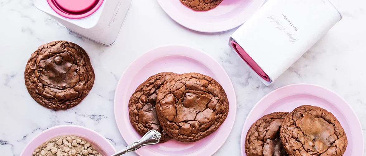 Ginger Elizabeth Chocolates chocolate truffle cookies on pink plates and classic hot chocolate tins on marble counter.