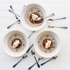 Ginger Elizabeth's Chocolate Molten Lava Cake. 3 individual servings in bowls  with  cream anglaise, whipped cream and toasted hazelnuts  surrounded by spoons on white bead board background .