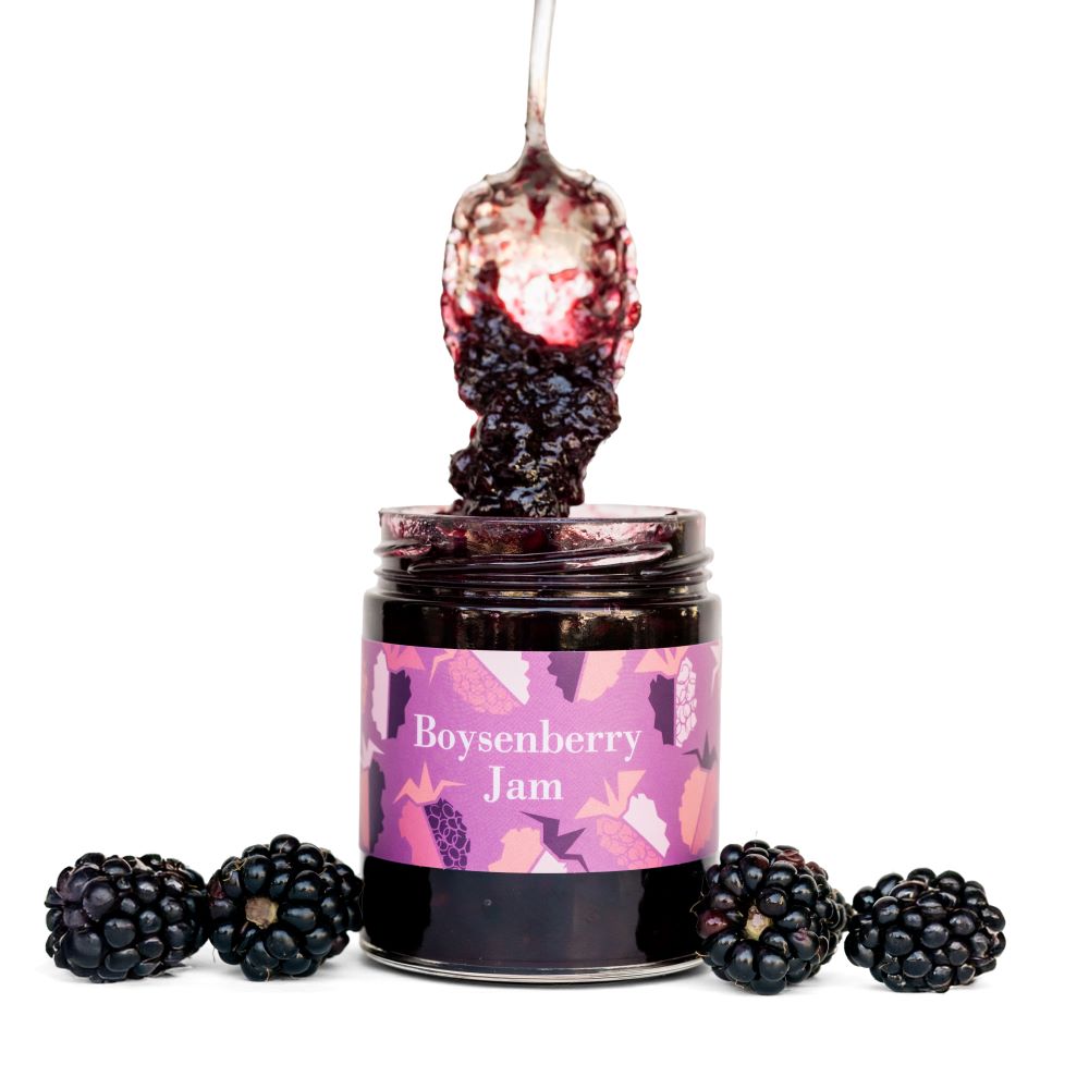 Jar of boysenberry jam with a spoonful of jam above it, surrounded by blackberries on a white background.