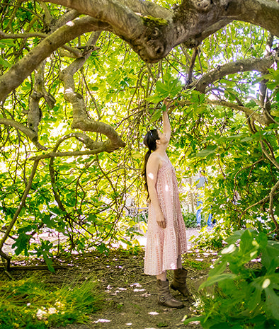 Ginger Elizabeth standing in right  side profile under trees reaching to branches above wearing pastel sundress and boots.