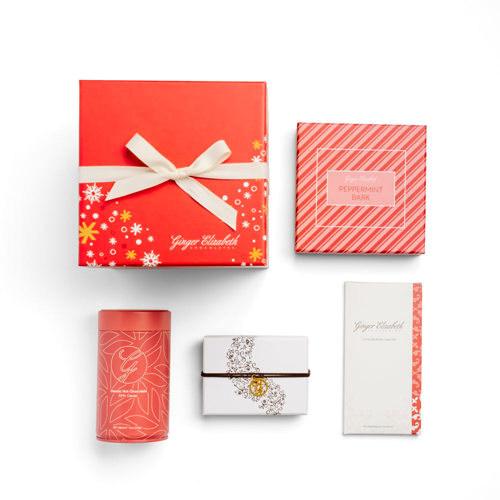 Ginger Elizabeth Chocolates holiday gift box with red lid with white and gold snowflakes and stars tied with white ribbon shown with box of peppermint bark, classic hot chocolate tin, classic 6 piece chocolate box and chocolate bar on white background.