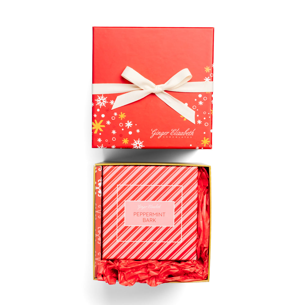 Ginger Elizabeth Chocolates holiday gift box with red lid with white and gold snowflakes and stars tied with white ribbon shown with open gift box with peppermint bark box surrounded by red tissue on white background.