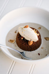 Ginger Elizabeth's Chocolate Molten Lava Cake. Individual servings in bowl with spoon,  cream anglaise, whipped cream and toasted hazelnuts on white bead board background .