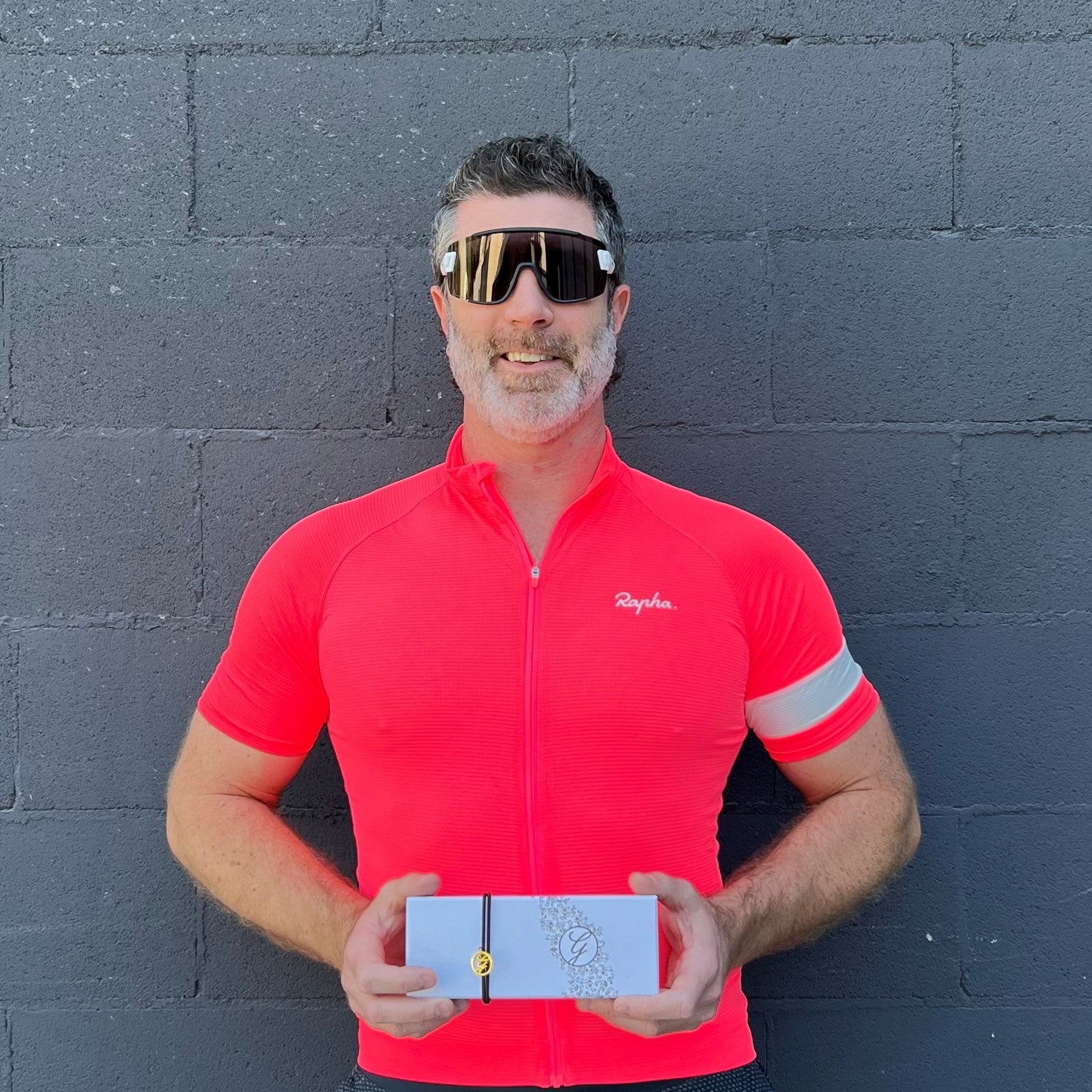 Man in red cycling jersey and sunglasses standing in front of painted brick wall holding 12pc chocolate box.