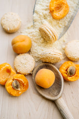 Ginger Elizabeth Chocolates Apricot Cobbler Macarons with Fresh apricots, flour and wooden spoon on light wood background