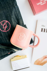Ginger Elizabeth Chocolates pink ceramic logo mug with folded apron and corners of macaron cookbook and pink candle  packaging in background 
