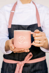 Ginger Elizabeth holding pink ceramic logo mug wearing denim apron with pink straps over chef whites top 