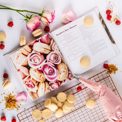 Ginger Elizabeth Chocolates Macaron Cookbook open to Raspberry Rose Macarons Page and surrounded by  roses and rose petals, macaron cookies, filled pastry bag and bakers cooling rack on white background