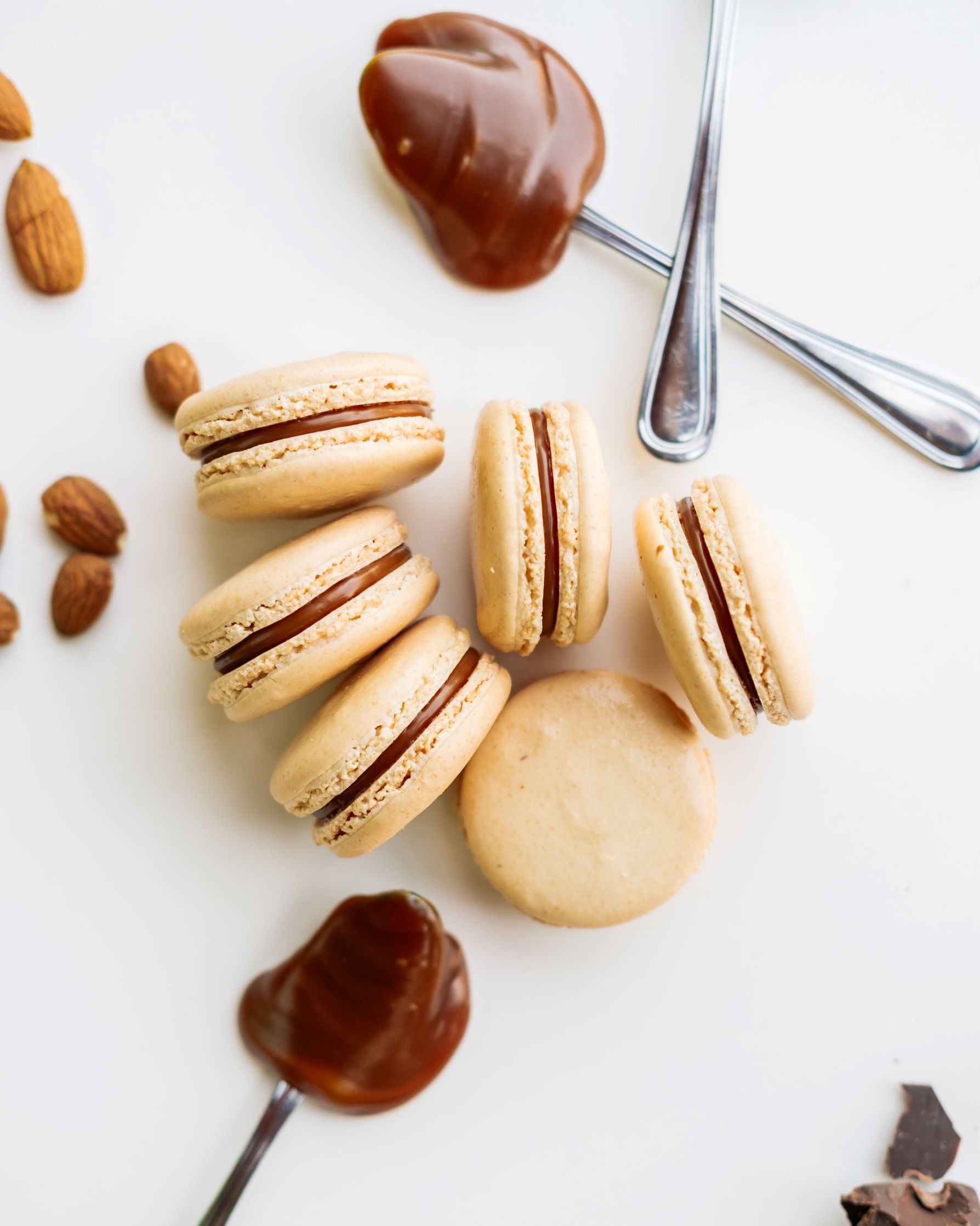 Six Salty Caramel Macarons shown with Caramel filling on spoons and scattering of fresh roasted almonds.