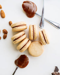 Six Salty Caramel Macarons shown with Caramel filling on spoons  and scattering of fresh roasted almonds on white background